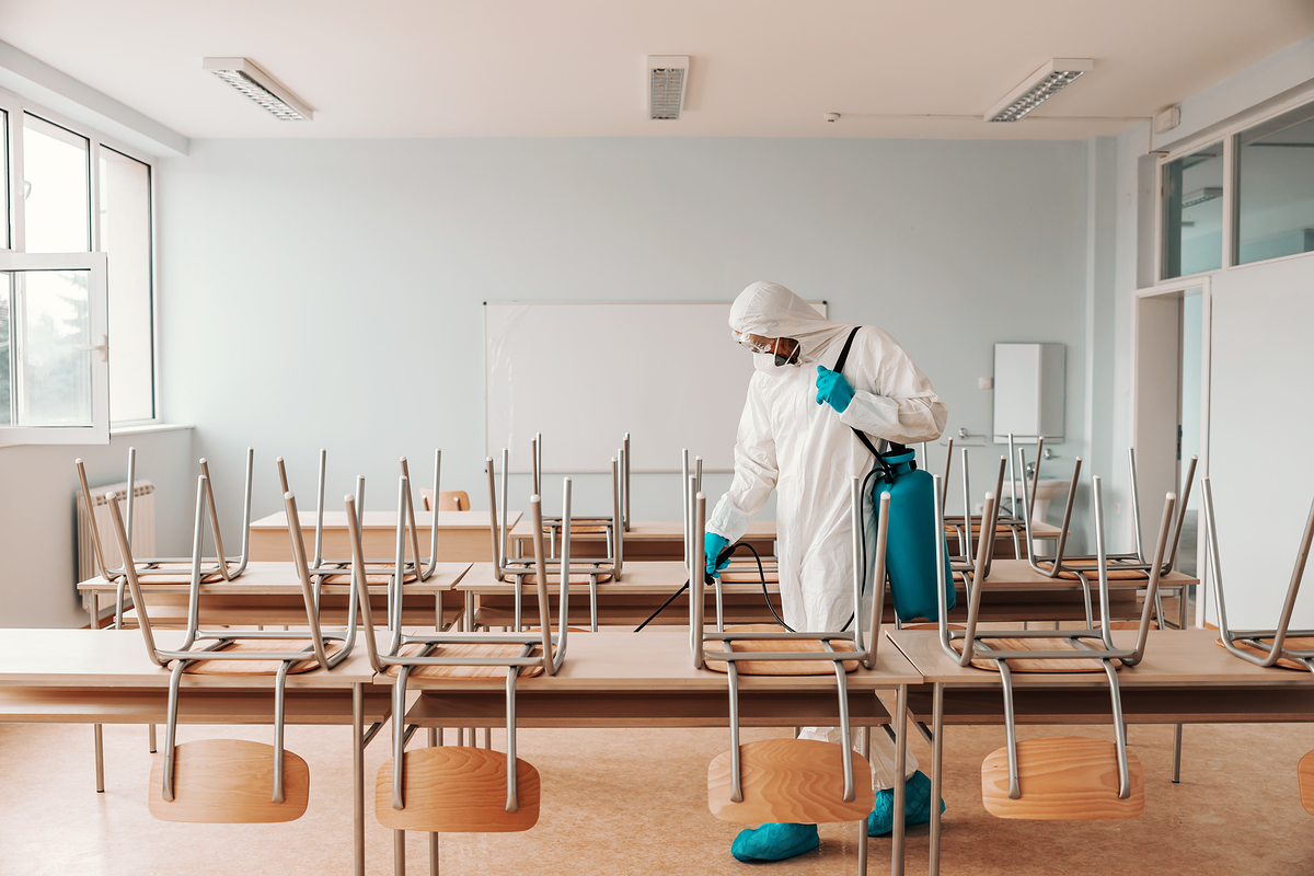 Man in sterile uniform, with gloves and mask holding sprayer and spraying with disinfectant floor in classroom.